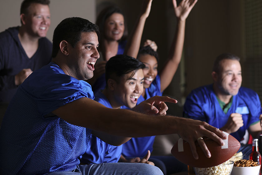 Multi-ethnic group of football fans watching the game together on television at home. They have snacks, drinks, and sports ball and wear blue team jerseys. Hispanic man foreground.