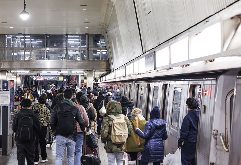 A large crowd of people waits on the platform and boards a subway train at Jamaica Station in Queens, New York City. The diverse group of commuters is dressed in winter clothing for the December weather. This scene captures the daily evening rush hour on the city's public mass transit system.
